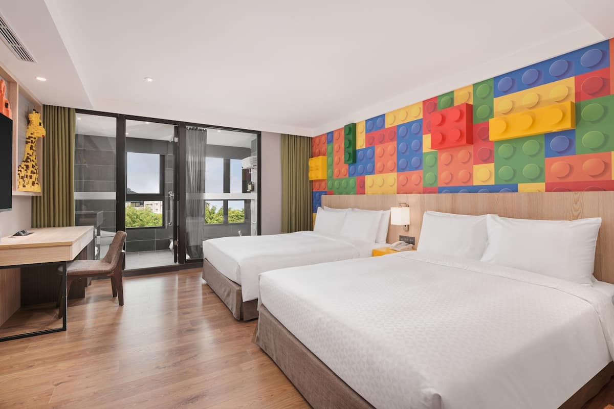 Quarto de hotel com duas camas, uma escrivaninha e vista através de uma porta de vidro.