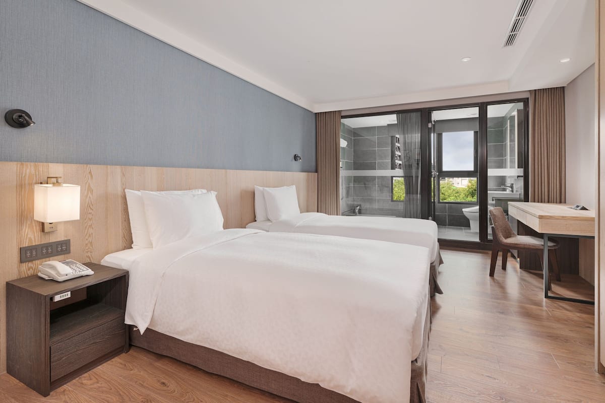 Quarto de hotel com duas camas, uma escrivaninha, uma cadeira, um telefone na mesinha de cabeceira e vista para o exterior pela porta de vidro.