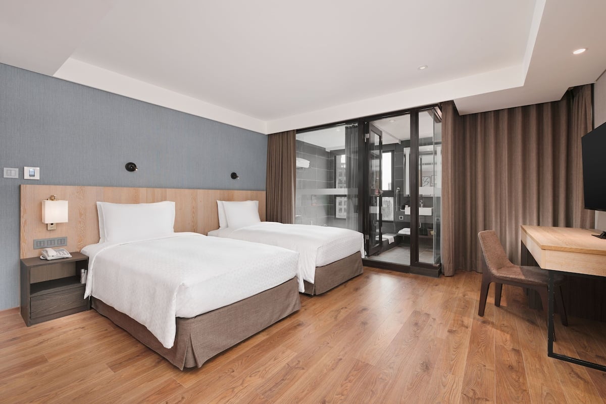 Quarto de hotel com duas camas, uma escrivaninha e vista da cidade através de uma grande porta de vidro.