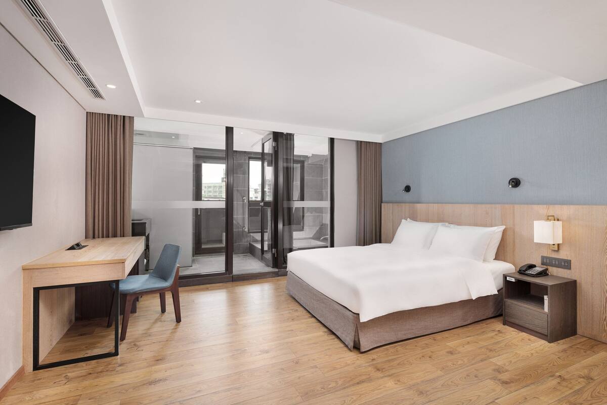 Quarto de hotel com cama, escrivaninha, cadeira e vista através de uma porta de vidro.