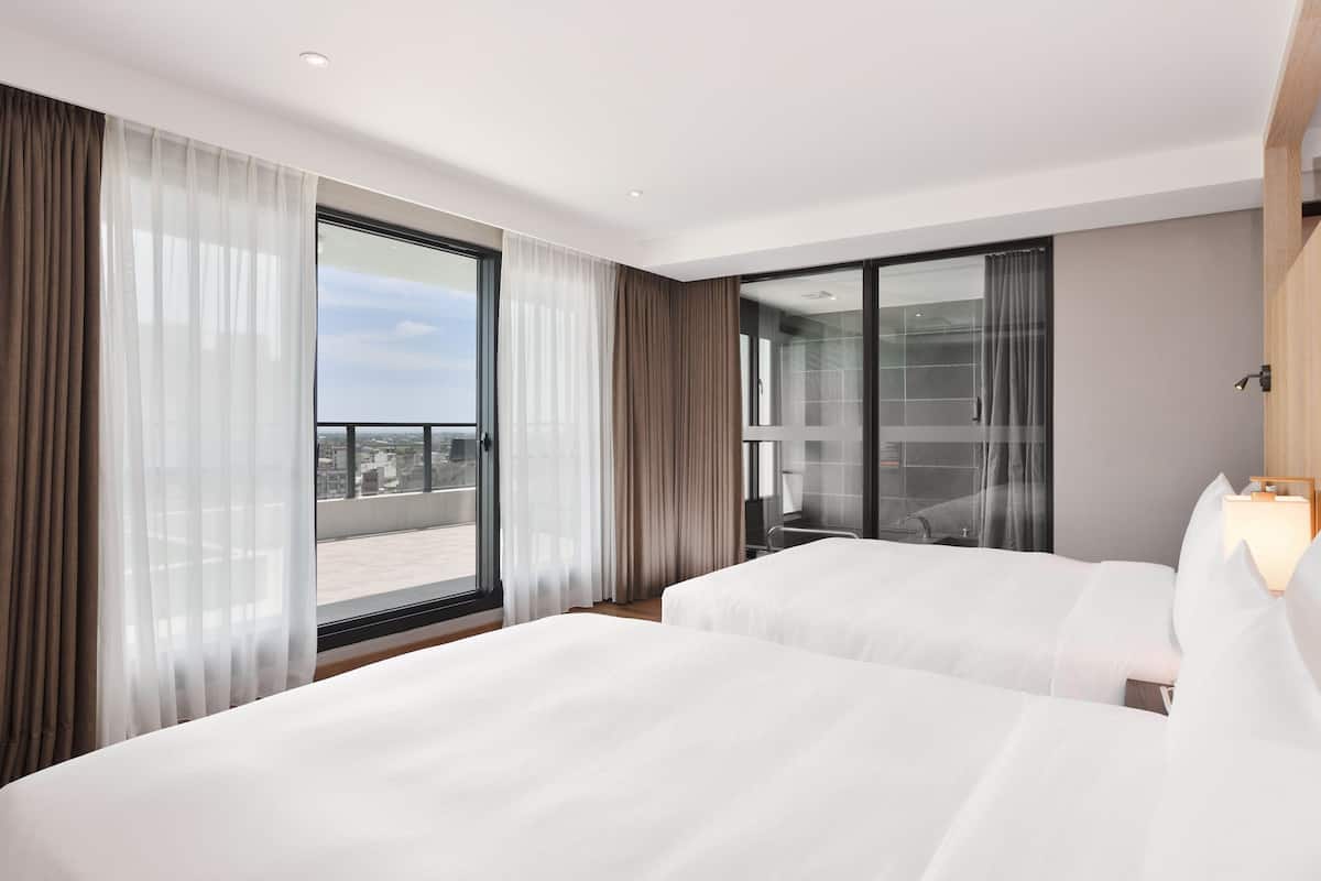 Quarto de hotel com duas camas, uma janela grande com cortinas leves, vista da cidade e uma varanda com porta de vidro.