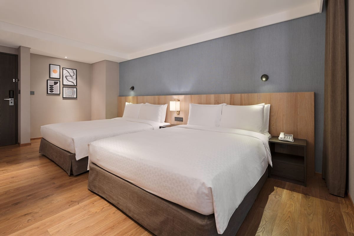 Quarto de hotel com duas camas, cabeceira de madeira, mesa de cabeceira e luminária na parede.