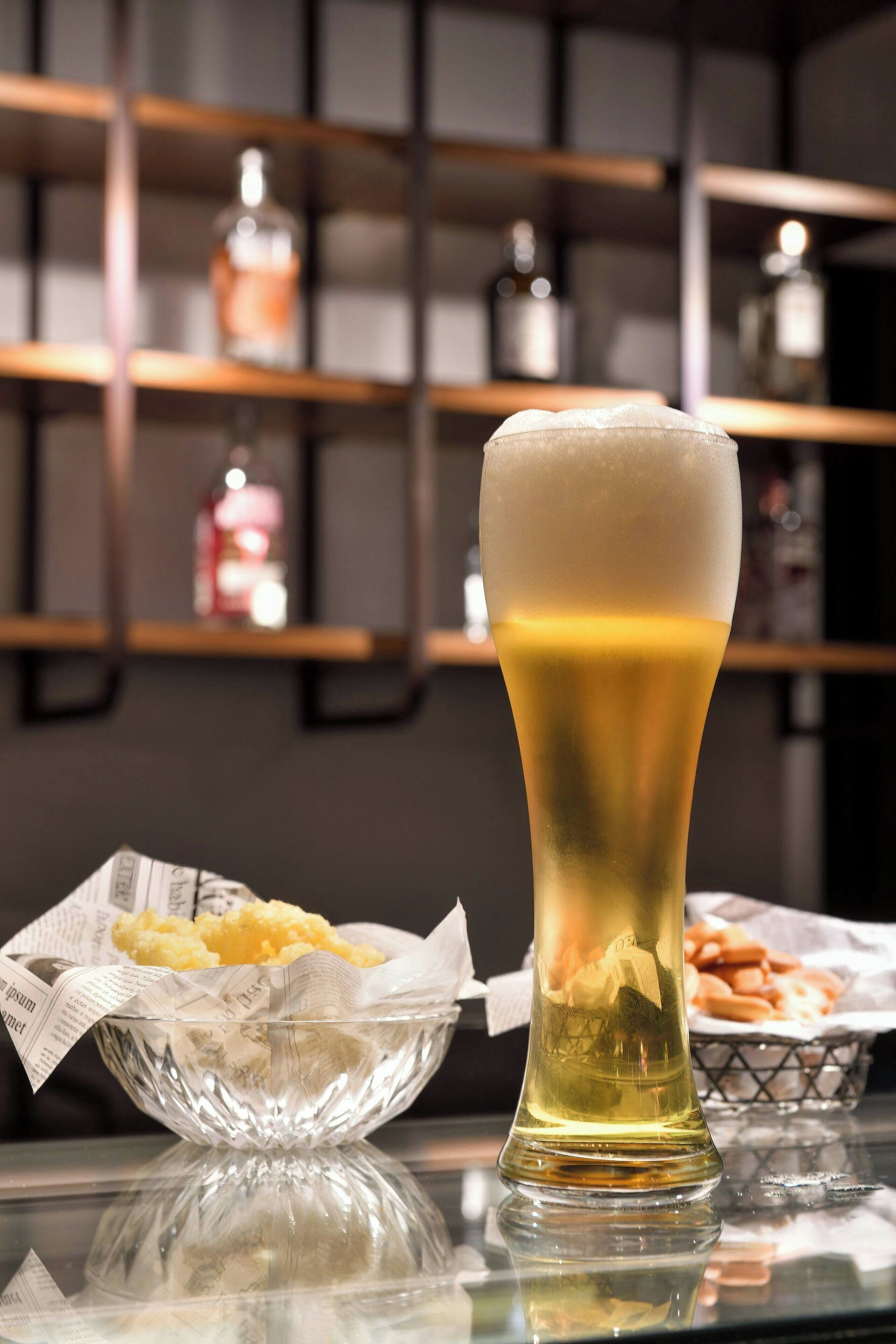 A tall glass of beer with a foamy head, placed on a reflective surface. In the background, there are shelves with bottles and a basket of fries.