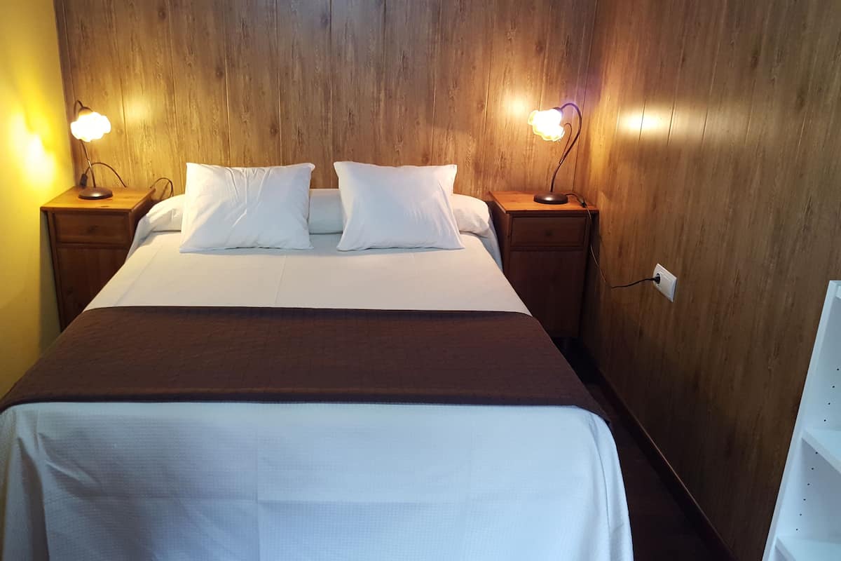 Una cama bien hecha con sábanas y almohadas blancas, flanqueada por dos mesitas de noche con lámparas, en una habitación con paneles de madera en las paredes.