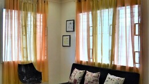 Una sala de estar con un sofá negro, una silla negra y un reposapiés estampado. Hay dos ventanas con cortinas translúcidas y una lámpara en el techo.