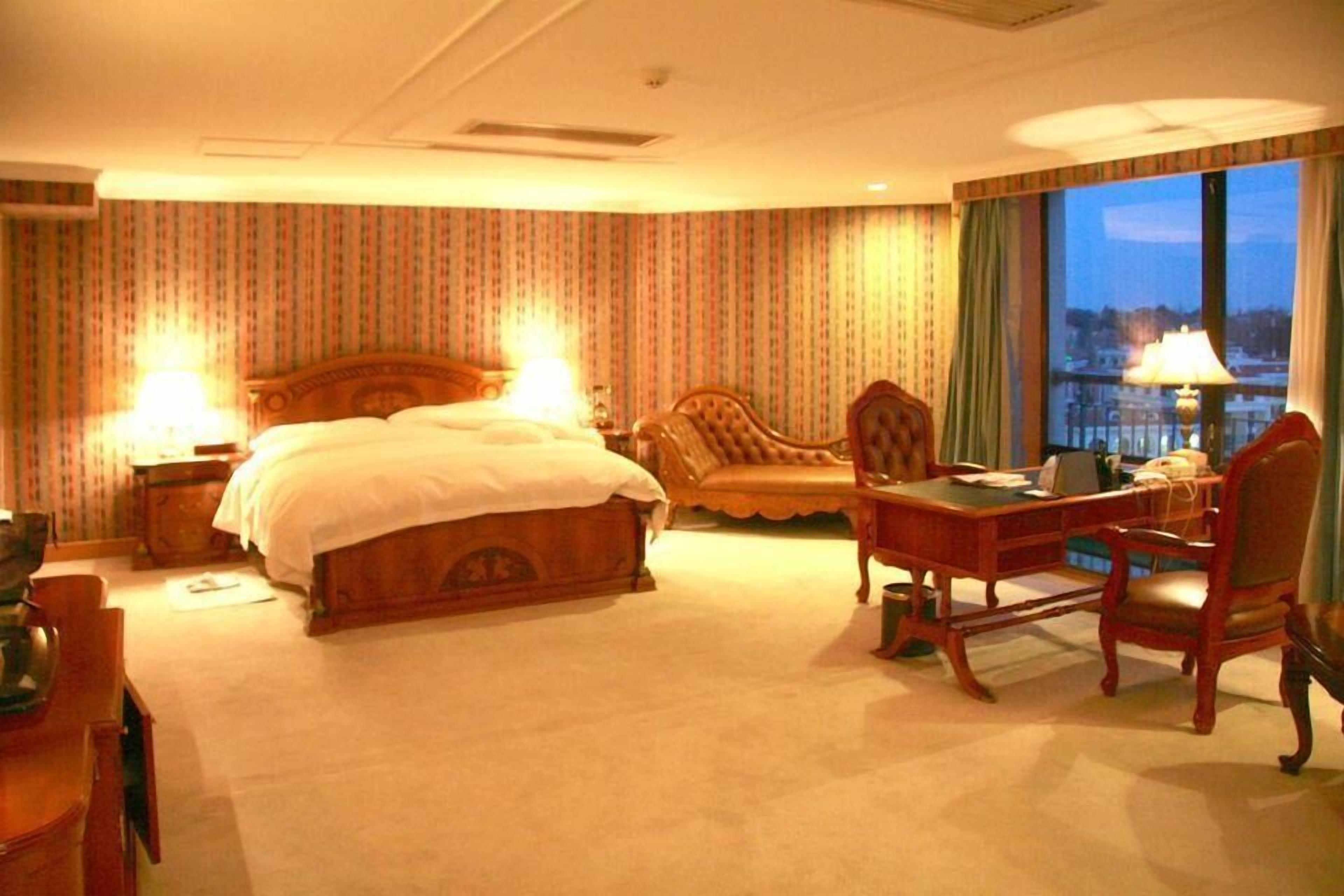 A hotel room with a large bed, a desk, a chair, and a view of the city through the window.