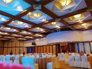 A large hall with a high ceiling featuring geometric patterns and recessed lighting. Tables are set up with white tablecloths and chairs, indicating a formal event or banquet setting.