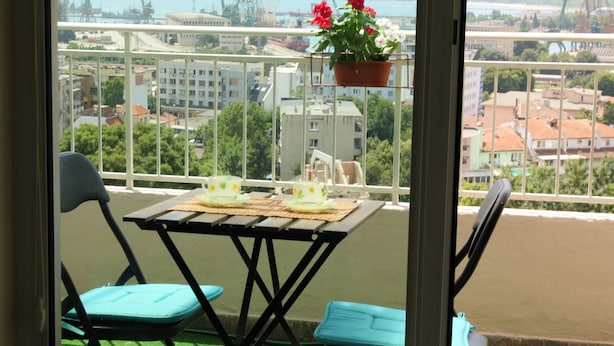 Un balcone con tavolo e sedie, un vaso di fiori appeso e vista su una città con edifici e un corpo d'acqua.