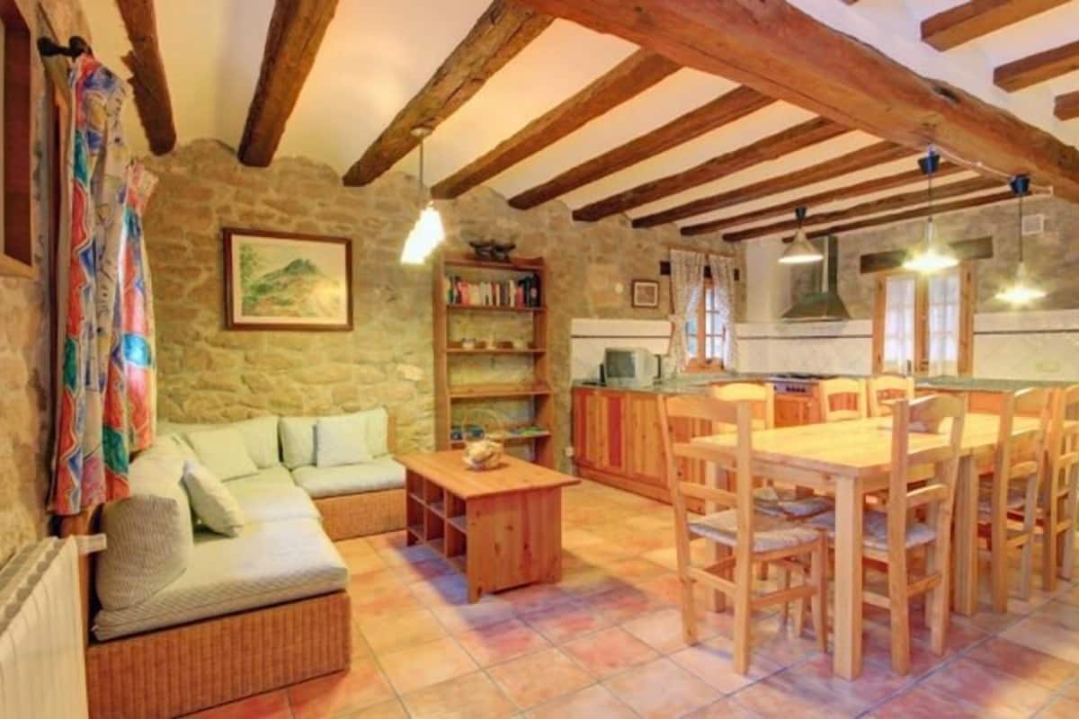 A cozy living room with a sofa, wooden dining table, and chairs, a kitchen area with a stove and cabinets, and a stone wall with a painting.