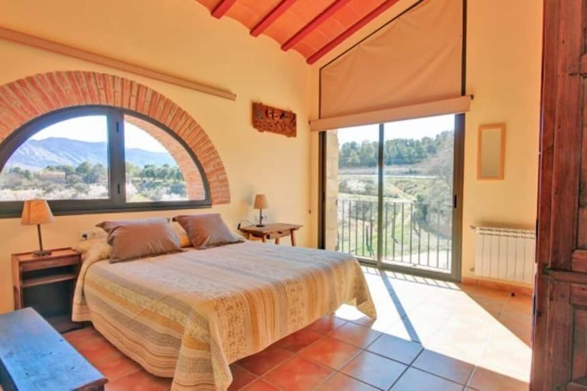 A bedroom with a large bed, a window with a view of the countryside, a wooden door, and a balcony.