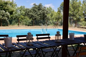 A poolside area with wooden tables and benches, a pool, and a view of trees and a fence in the background. - Mas del Cigarrer (Horta de Sant Joan)