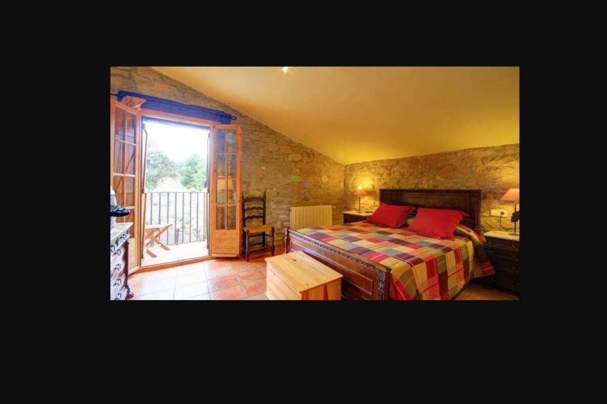 A bedroom with a stone wall, a bed with a colorful quilt, a wooden bench, a nightstand, and a window with a view of greenery.