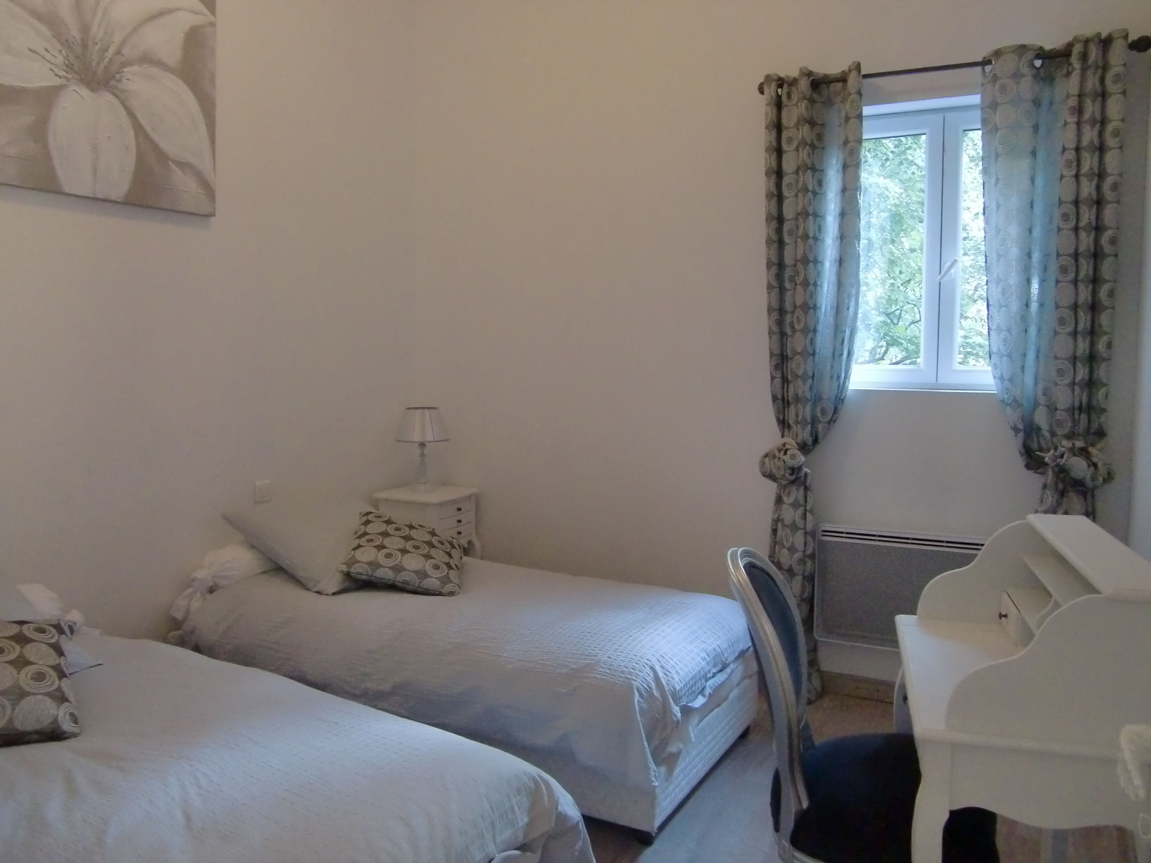 A bedroom with two beds, a bedside table with a lamp, a chair, and a window with curtains.