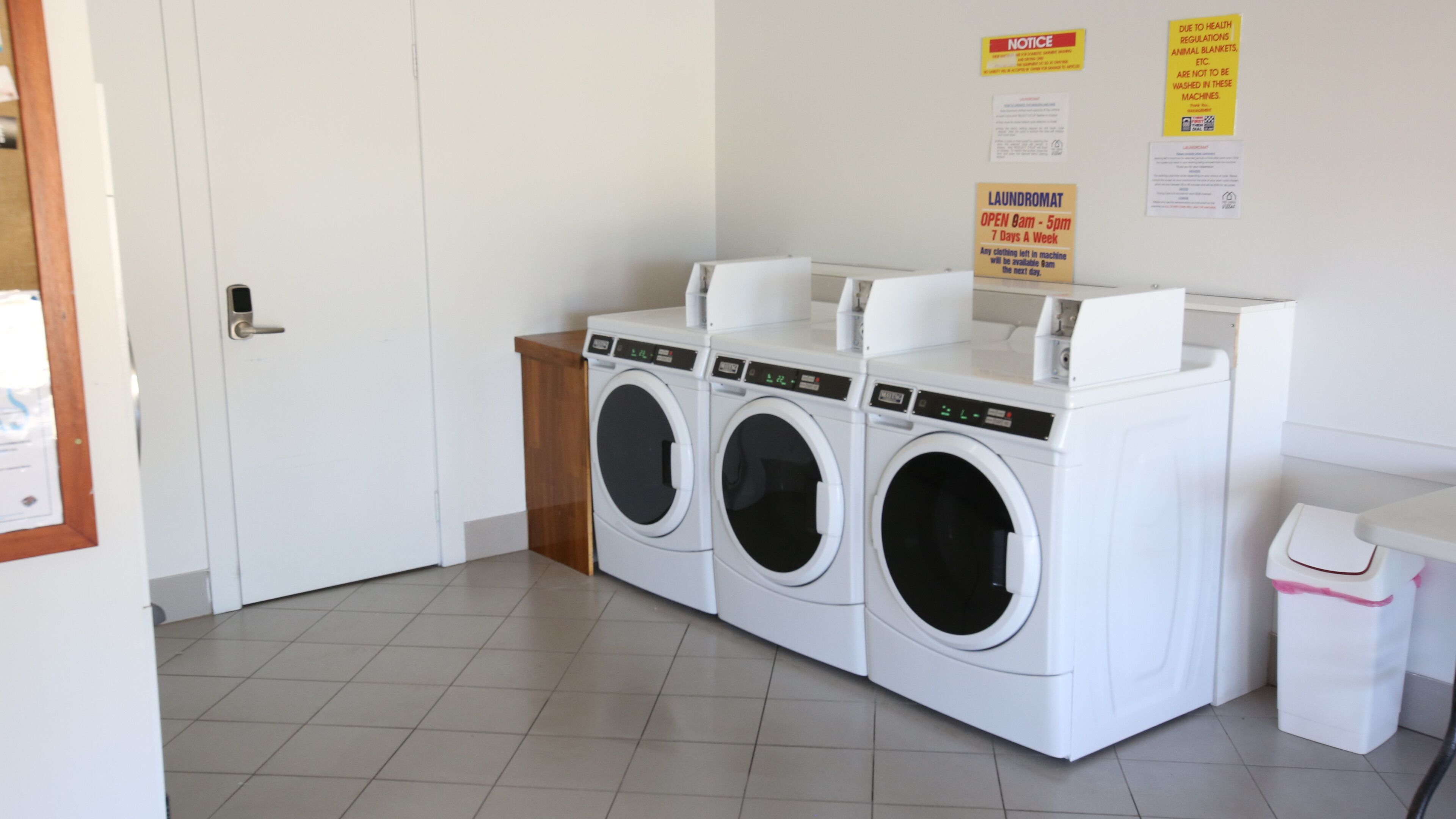 A laundry room with three stacked washing machines, a notice board with health regulations, and a door.