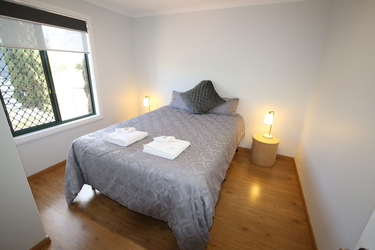 Una cama bien hecha con una colcha gris estampada, dos almohadas y dos toallas blancas dobladas. Hay dos lámparas de noche sobre mesitas de madera.