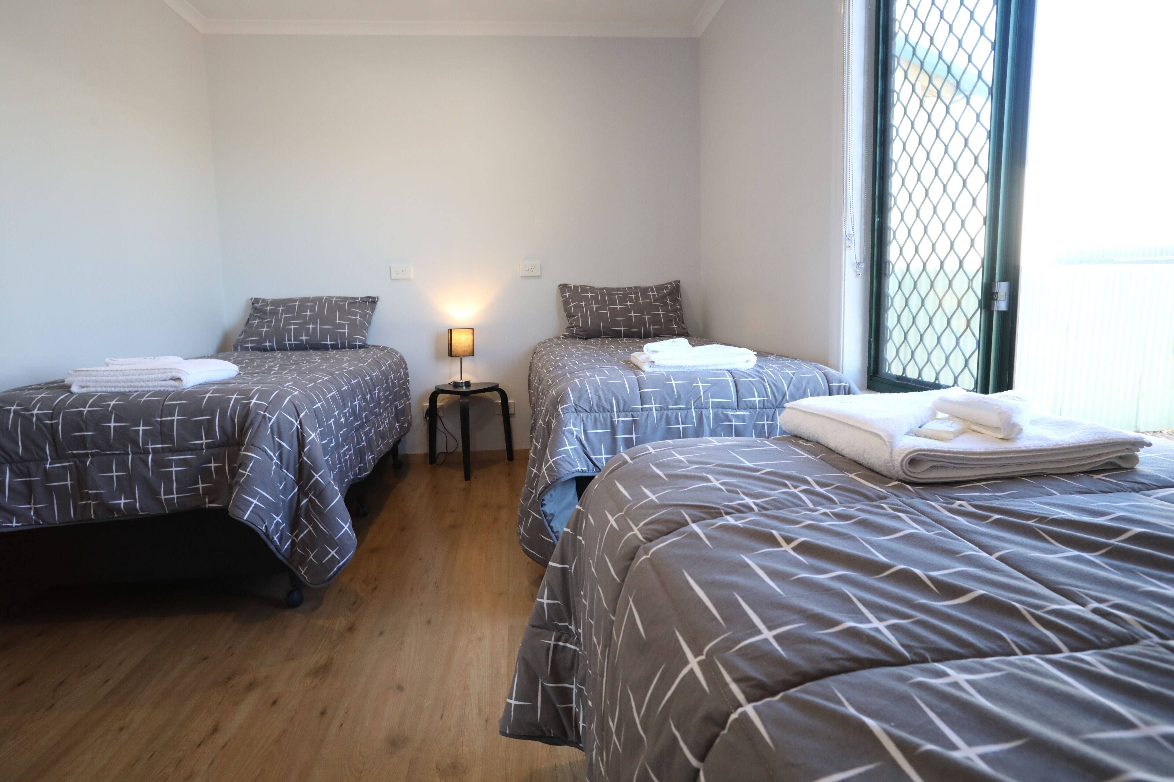 Een hotelkamer met twee bedden, elk met een beddekleed en kussens met een patroon, een klein nachtkastje met een lamp en een raam met hor.