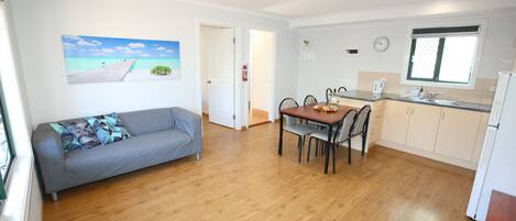 A modern kitchen with wooden flooring, a dining table with chairs, a grey sofa, and a painting of a pier.