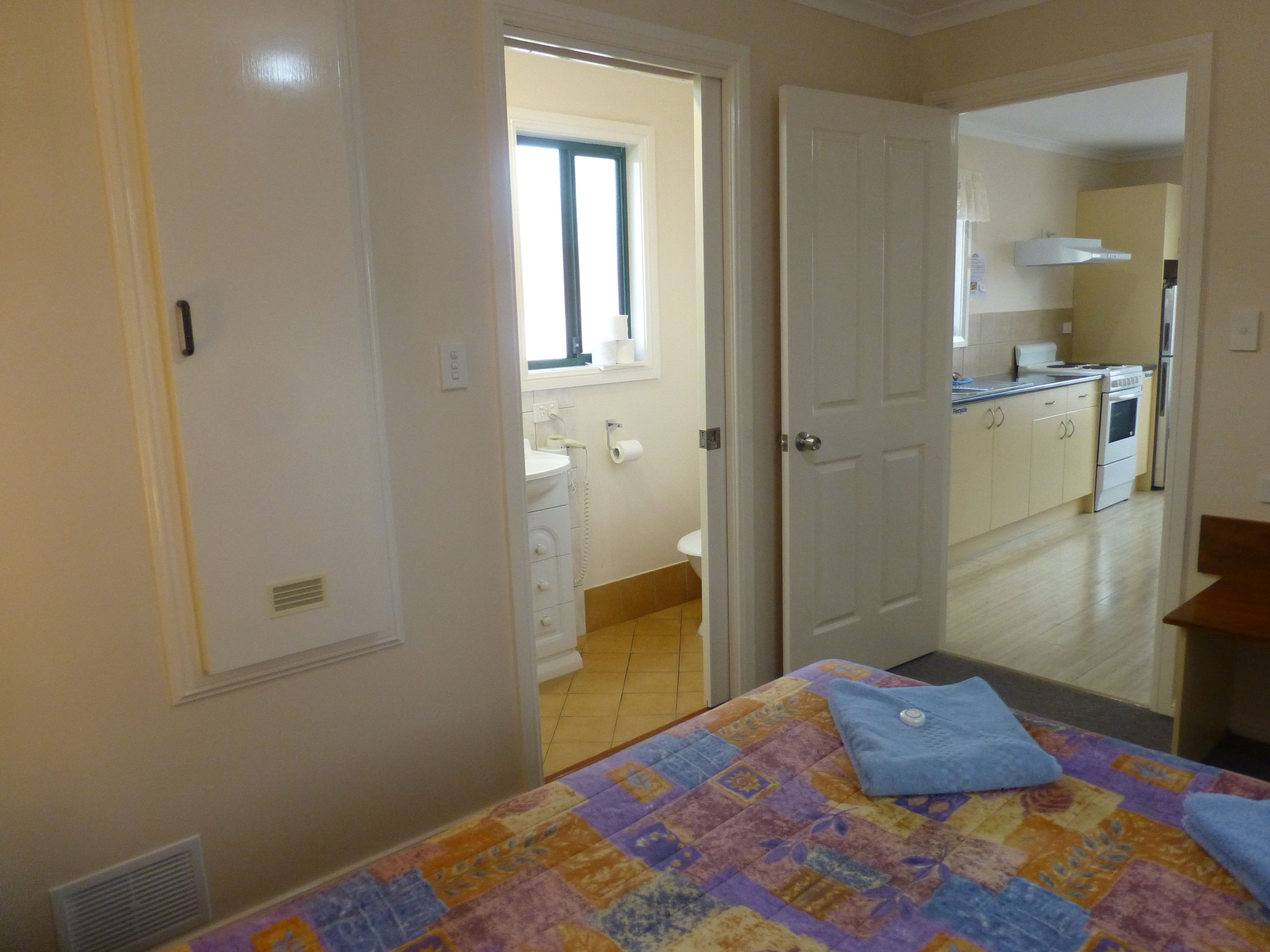 Un dormitorio con cama, un baño a la vista a través de una puerta abierta y una cocina al fondo.
