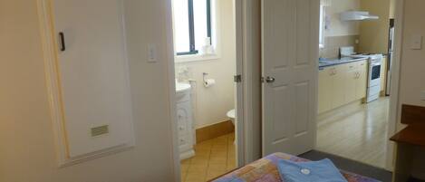 Un dormitorio con cama, un baño a la vista a través de una puerta abierta y una cocina al fondo.