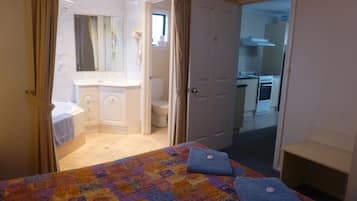 Une chambre avec un lit, une salle de bain avec un lavabo et des toilettes, et une cuisine visible à travers une porte ouverte.