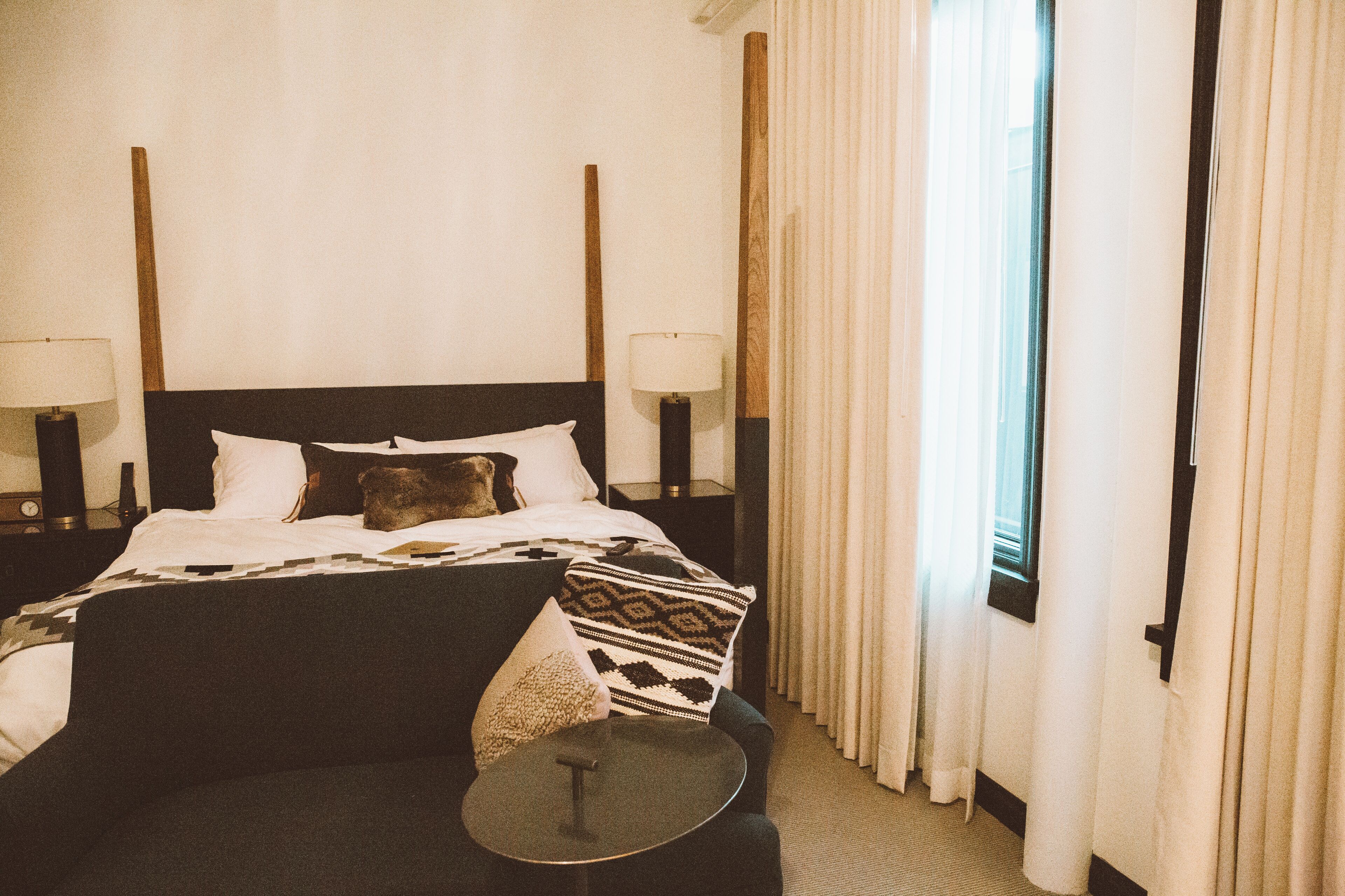 Quarto de hotel com uma cama grande, duas mesas de cabeceira com luminárias, uma mesa redonda pequena e uma janela com cortinas.