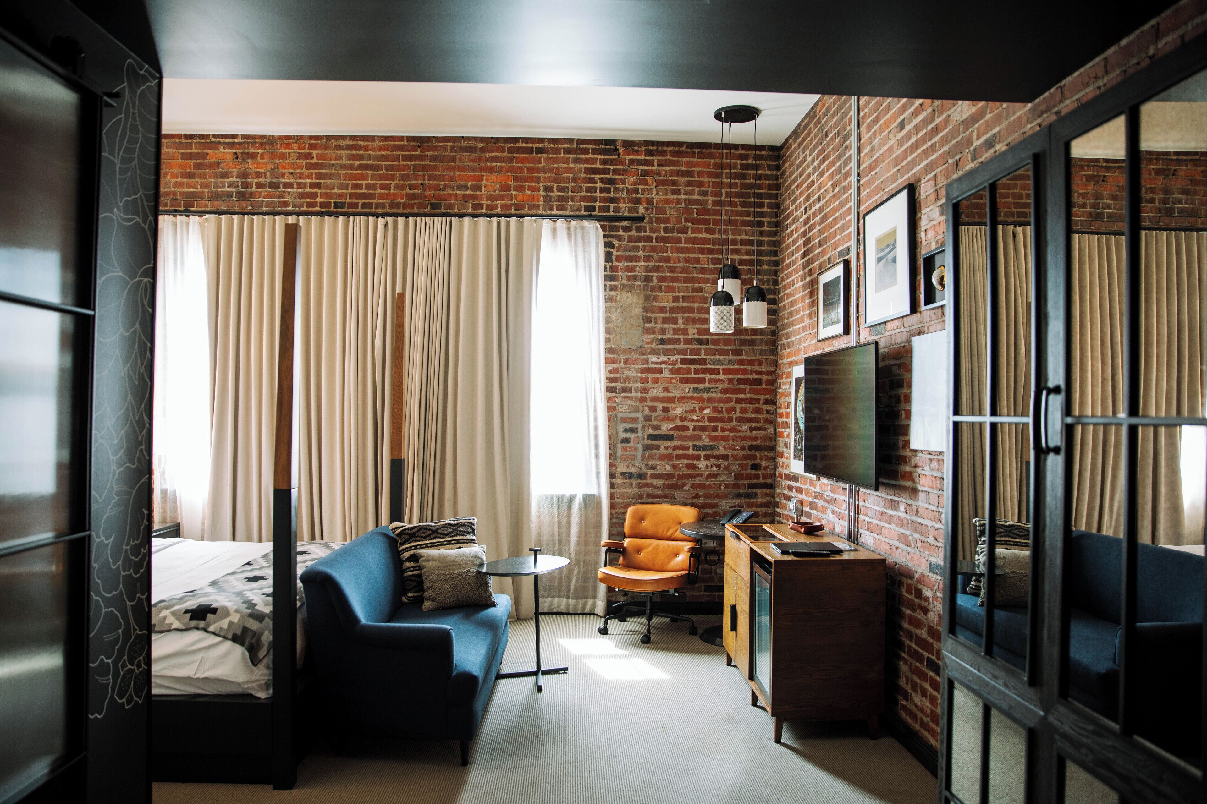 A compact hotel room with a brick wall, a bed with a patterned bedspread, a blue sofa, a small round table, an orange chair, a wooden cabinet, and a television mounted on the wall.