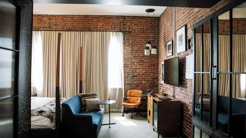 A compact hotel room with a brick wall, a bed with a patterned bedspread, a blue sofa, a small round table, an orange chair, a wooden cabinet, and a television mounted on the wall.