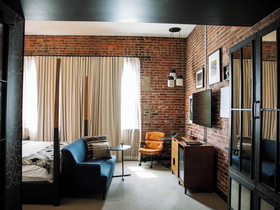 A compact hotel room with a brick wall, a bed with a patterned bedspread, a blue sofa, a small round table, an orange chair, a wooden cabinet, and a television mounted on the wall.