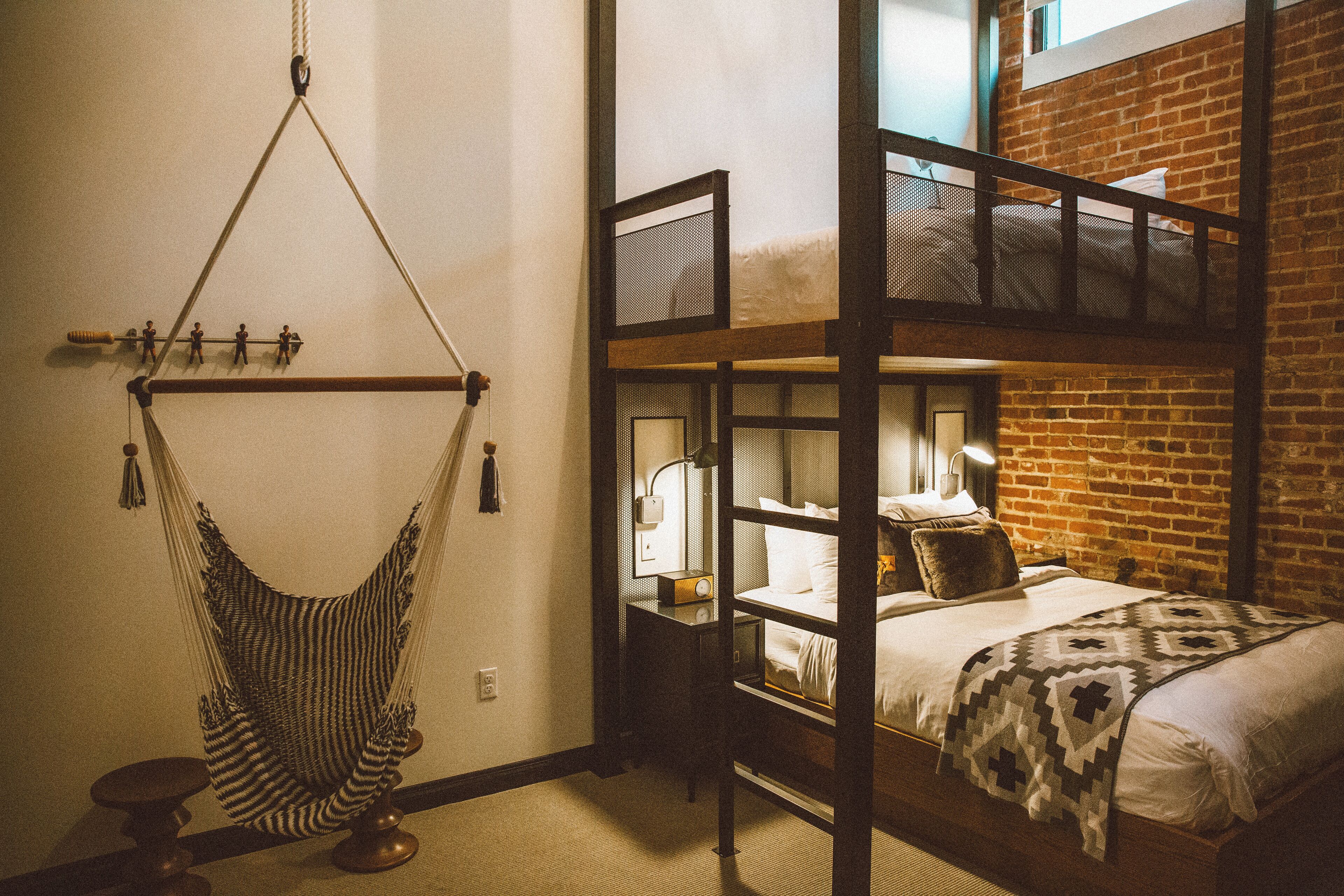 Una habitación con hamaca, litera, mesita de noche con lámpara y pared de ladrillo.