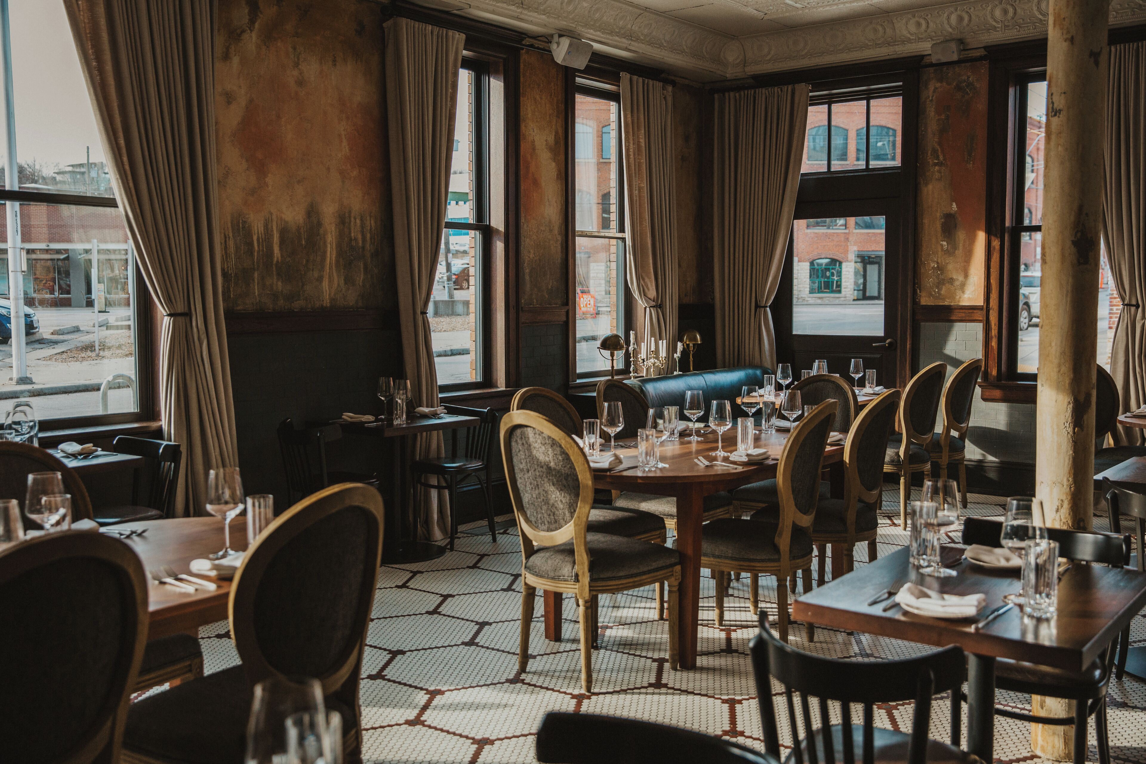 Un restaurante con una decoración clásica, con mesas y sillas de madera, ventanas amplias con cortinas y vista a la calle.