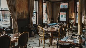 Un restaurante con una decoración clásica, con mesas y sillas de madera, ventanas amplias con cortinas y vista a la calle.