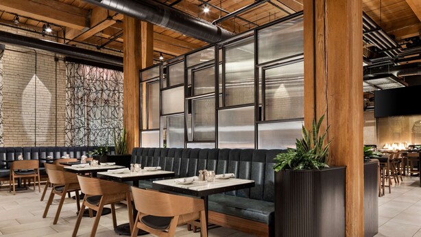 L’intérieur d’un restaurant moderne, avec des poutres en bois, un coin salon capitonné noir et une grande fenêtre aux montures métalliques.