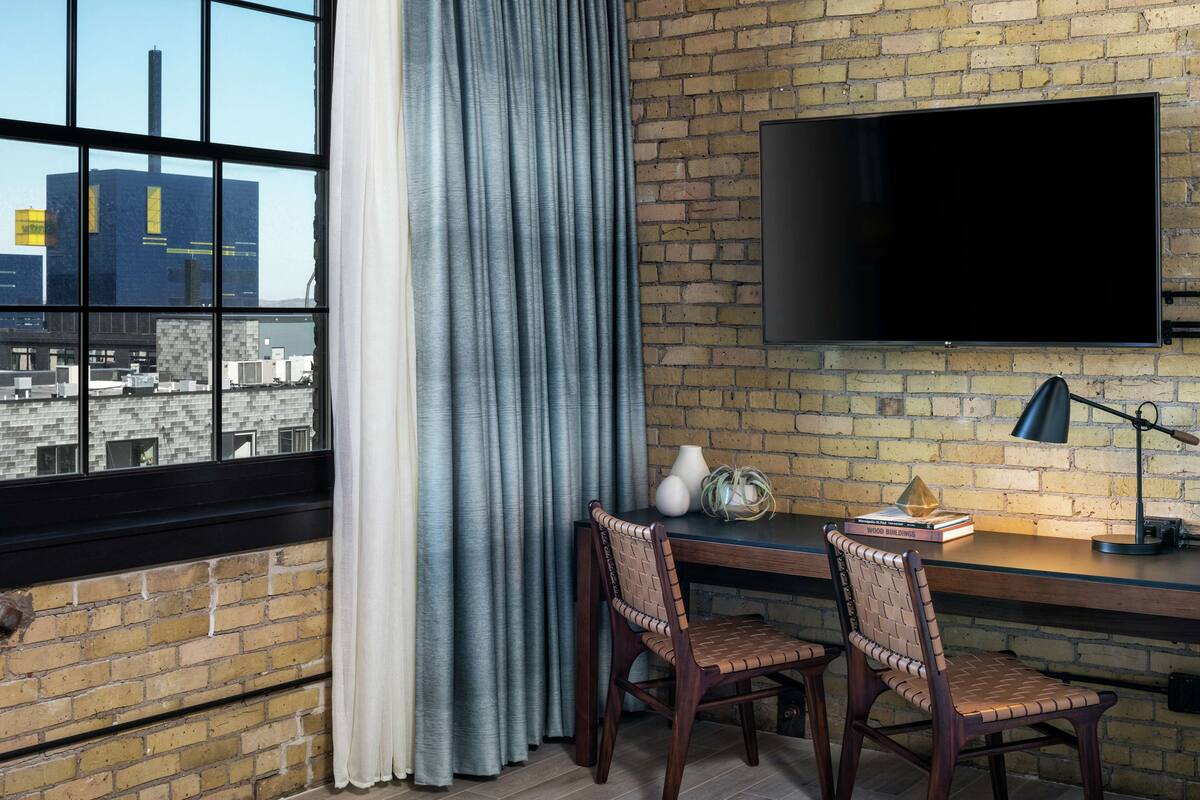 A modern room with a brick wall, a large window with curtains, a flat-screen TV, two wooden chairs, a small table with books and a lamp, and a cityscape view.