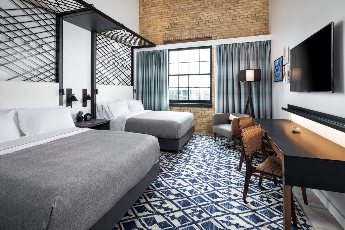 A hotel room with two beds, a desk, a chair, a TV, and a large window with curtains.