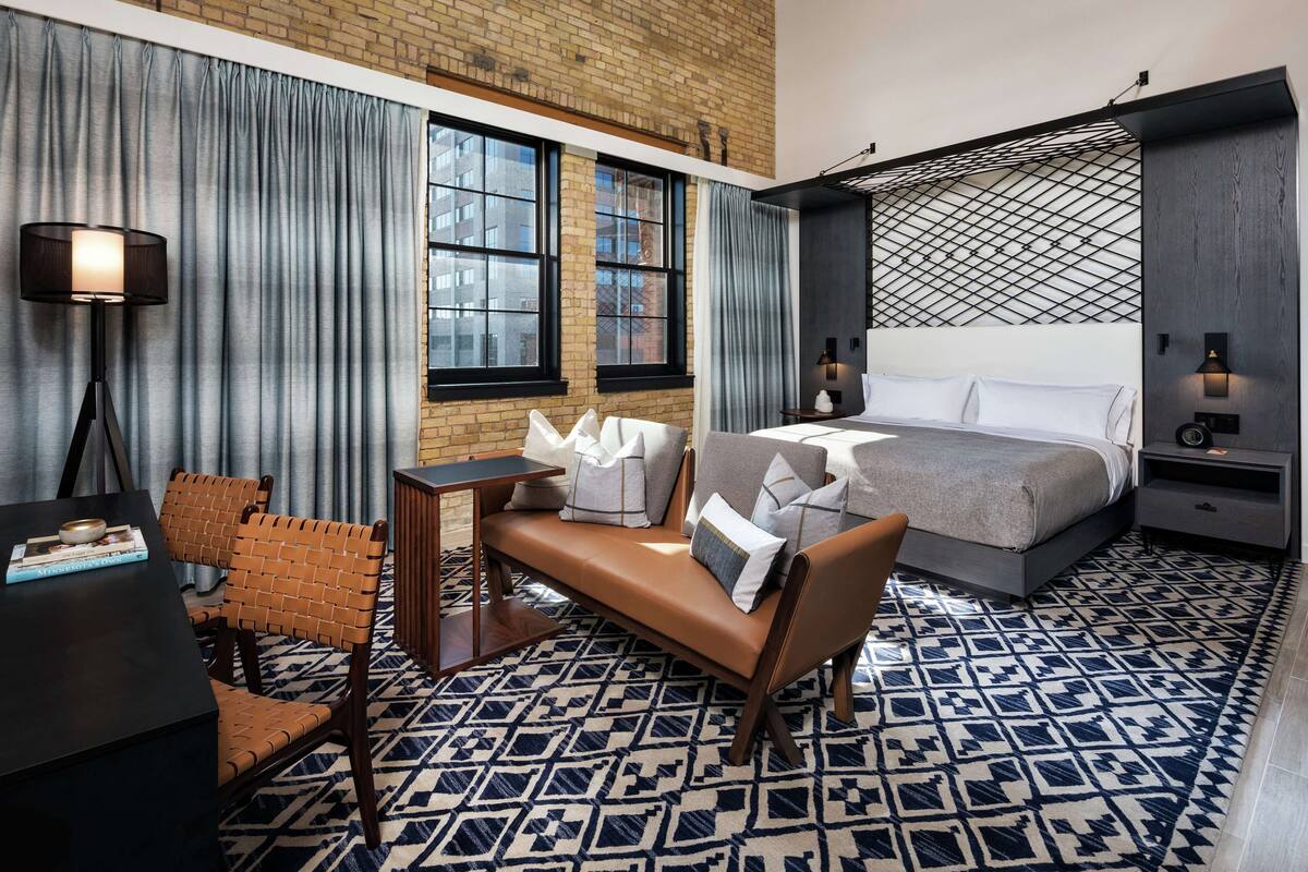 A modern hotel room with a brick wall, large windows, a patterned floor, a bed, a desk, and a chair.