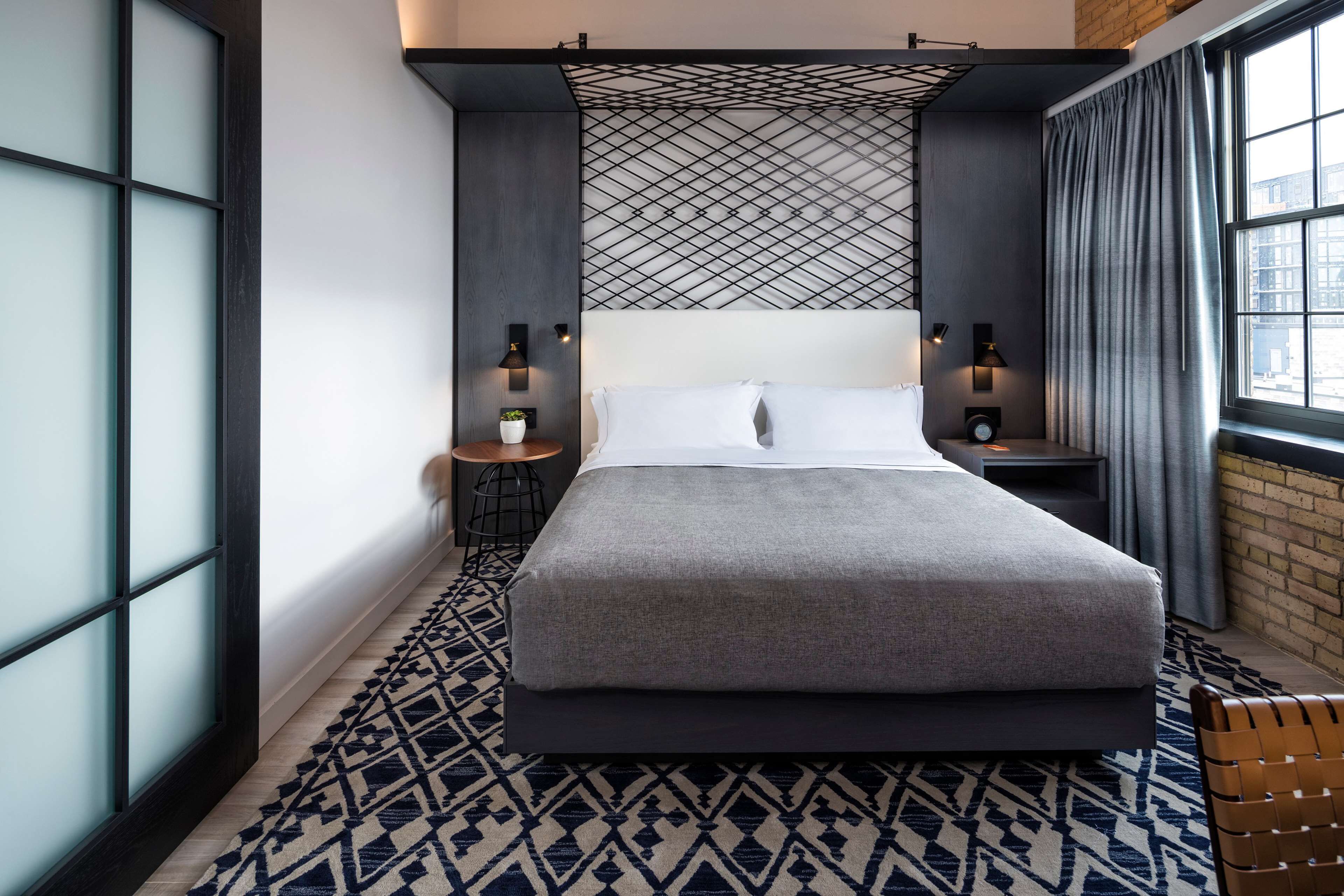 A modern hotel room with a large bed, a patterned rug, a small side table with a plant, and a window with curtains.