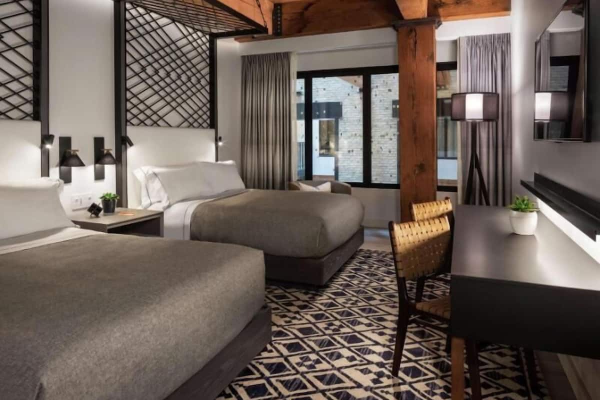 A hotel room with two beds, a desk, a chair, a large window, and a patterned floor.