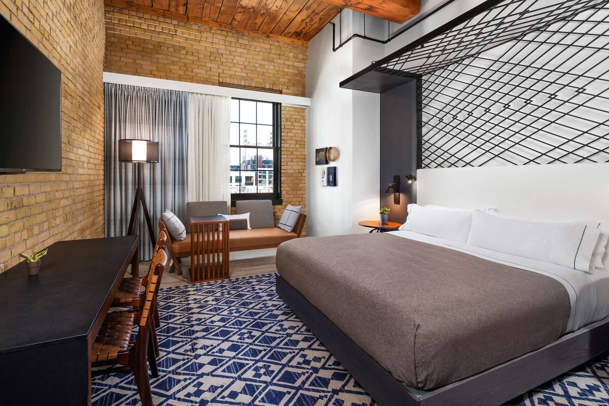 A modern hotel room with a brick wall, a large bed, a desk, and a view of the city through a window.