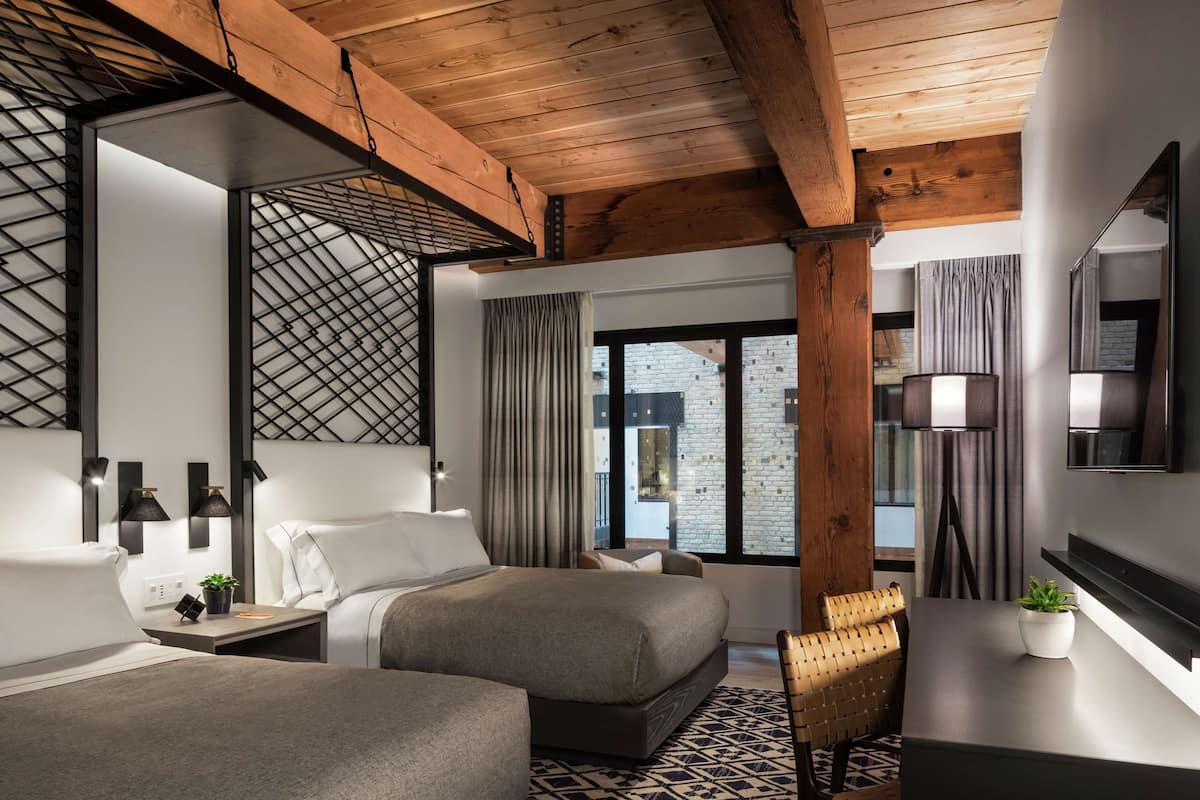 A modern hotel room with two beds, a desk, a chair, and a large window with curtains.