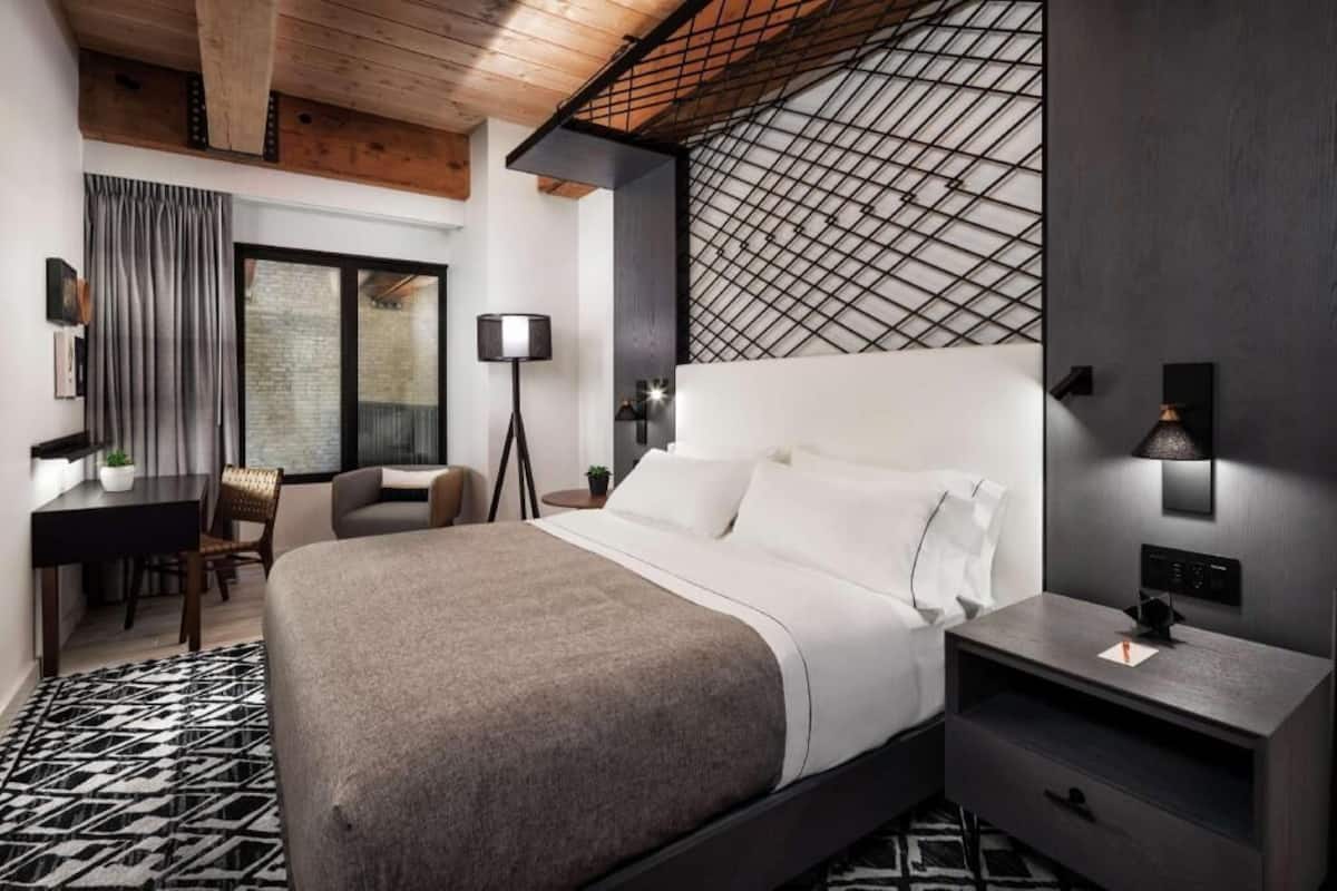 A modern hotel room with a large bed, a desk, a chair, and a patterned rug.