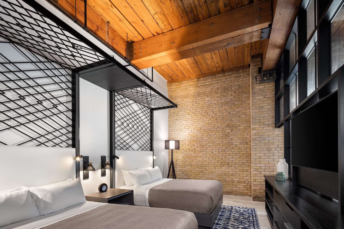A modern hotel room with a brick wall, wooden ceiling beams, a large flat-screen TV, two beds with white linens, a patterned rug, and a nightstand with a lamp.