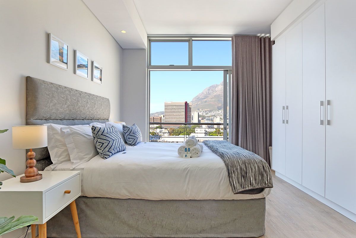 Ein modernes Hotelzimmer mit einem großen Bett, einem Nachttisch mit Lampe, einem Blick auf die Stadt durch ein großes Fenster und einem Kleiderschrank.