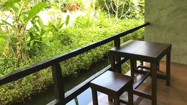 Una mesa y banco de madera junto a una ventana con vista a un jardín exuberante, con diversas plantas y una pequeña fuente.