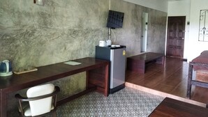 Una habitación con un escritorio de madera, una silla blanca, un refrigerador, un televisor montado en la pared y una alfombra estampada.