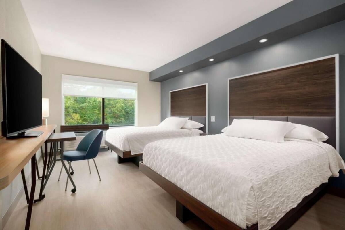 Une chambre d’hôtel avec deux lits, un bureau avec une chaise, une télévision et une fenêtre donnant sur un paysage verdoyant.