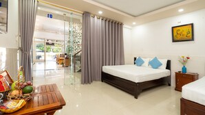 A modern hotel room with a large bed, a wooden table with fruits and a basket, a small bedside table with a vase, and a view of the outdoor area through a glass door. - The Art - Big Sea Villa Hoi An Riverside (Da Nang)
