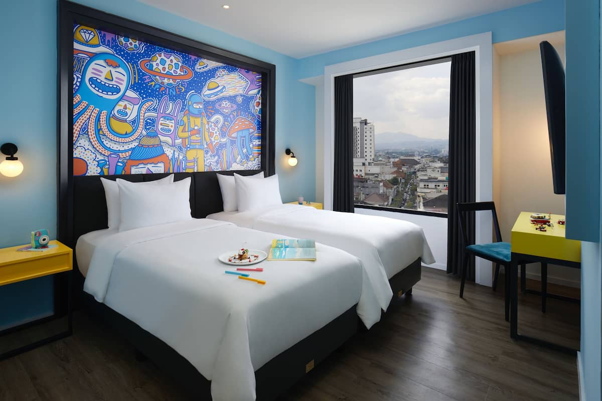 Habitación de hotel con dos camas, un ventanal con vista a la ciudad, un cuadro abstracto colorido en la pared y una mesita con un juguete.