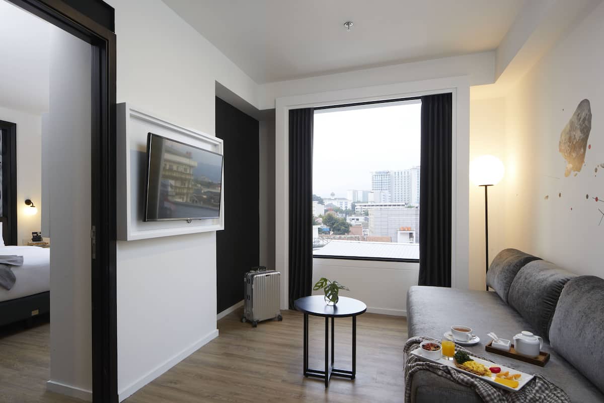 Una habitación de hotel moderna con una cama, un sofá, una pequeña mesa con desayuno y vistas a la ciudad a través de amplias ventanas.