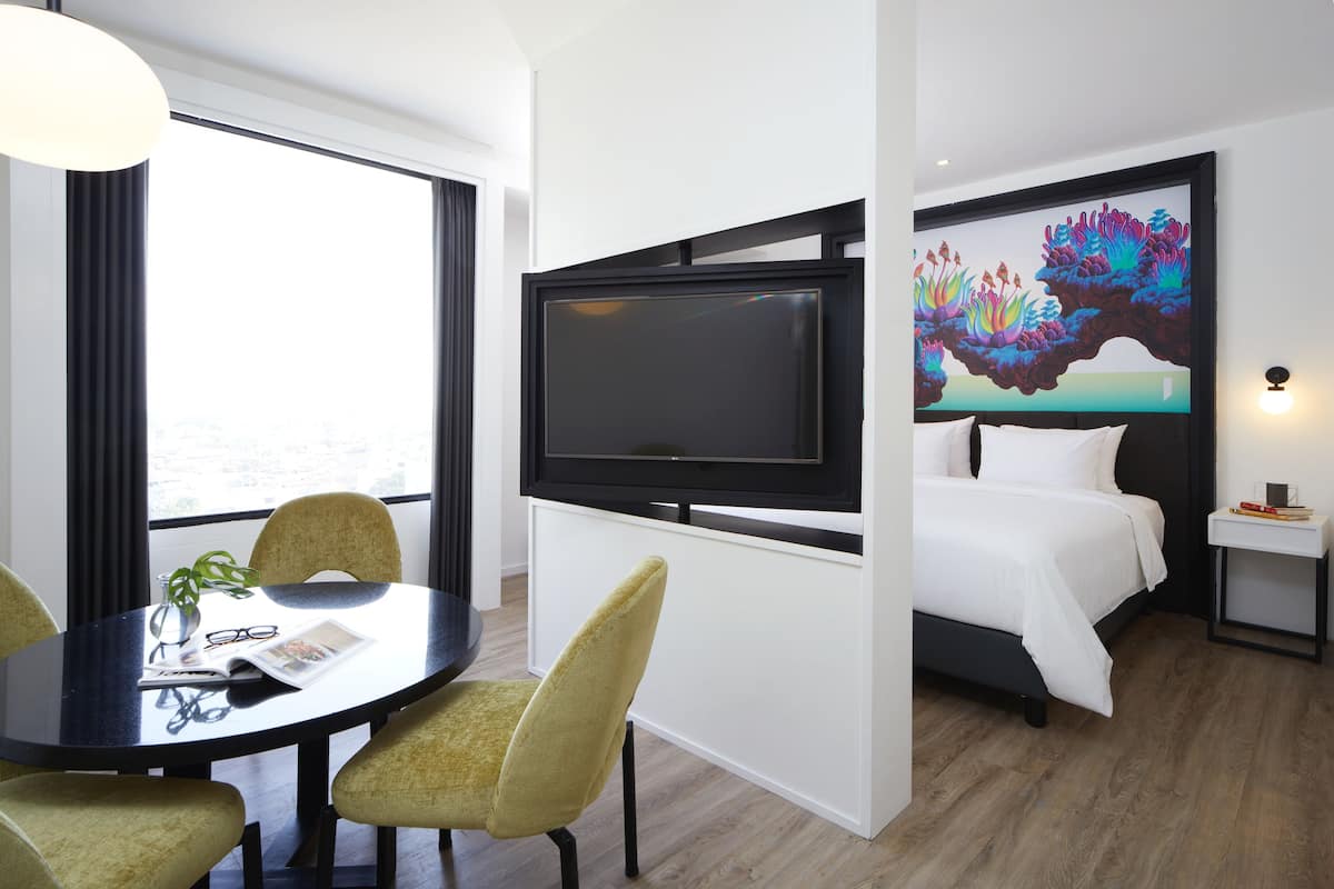 Habitación de hotel con cama, televisión, mesa de comedor y sillas, y un ventanal grande.