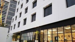 A modern building with a white facade, multiple windows, and a glass entrance area with chairs and tables. - de Braga, ARTOTEL Curated (Bandung)